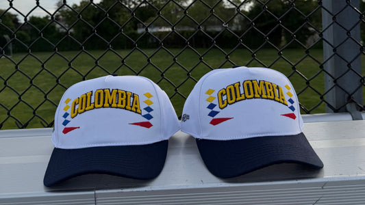 Two white and navy blue baseball caps with 'COLOMBIA' embroidered in yellow on the front, resting on a white bench in front of a chain-link fence at a baseball field.